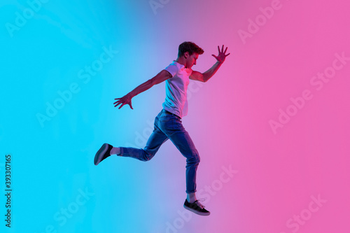 Running on. Young caucasian man's jumping high on gradient blue-pink studio background in neon light. Concept of youth, human emotions, facial expression, sales, ad. Beautiful model in casual.