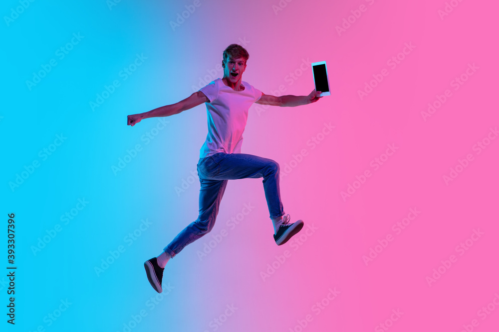 Winner with tablet. Young caucasian man's jumping high on gradient blue-pink studio background in neon light. Concept of youth, human emotions, facial expression, sales, ad. Beautiful model in casual.