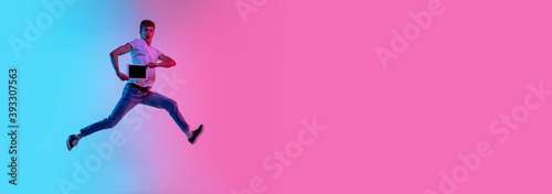 Jumping high with tablet  flyer. Young man s portrait on gradient blue-pink studio background in neon light. Concept of youth  human emotions  facial expression  sales  ad. Beautiful model in casual.