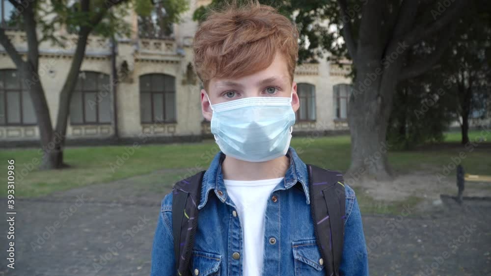 Little redhead boy putting on Covid-19 face mask and looking at camera. Portrait of Caucasian schoolboy posing outdoors during coronavirus viral pandemic. Covid lifestyle concept. Little redhead boy putting on Covid-19 face mask and looking at camera. Portrait of Caucasian schoolboy posing outdoors during coronavirus viral pandemic. Covid lifestyle concept.