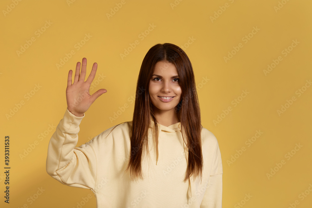 Open to communication. Cheerful friendly caucasian woman in yellow hoodie waves palm in hello gesture, cute toothy smiles. isolated over a yellow background. Open to communication. Cheerful friendly caucasian woman in yellow hoodie waves palm in hello gesture, cute toothy smiles. isolated over a yellow background.
