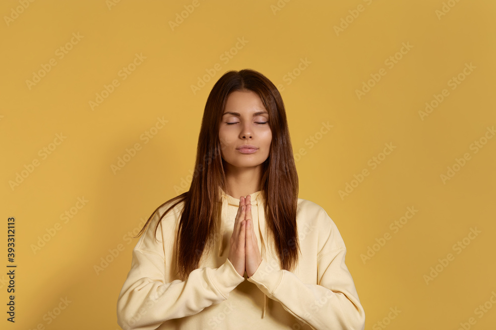 Cheerful caucasian woman in yellow hoodie, holding hands in namaste or prayer, keeping eyes closed while practising yoga and meditating, have calm look on her face. Isolated over a yellow background. Cheerful caucasian woman in yellow hoodie, holding hands in namaste or prayer, keeping eyes closed while practising yoga and meditating, have calm look on her face. Isolated over a yellow background.