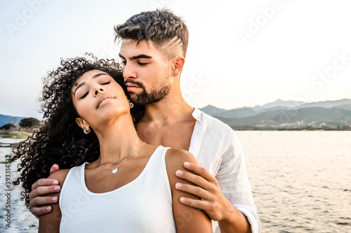 Heterosexual mixed race couple in romance scene at sunset with vintage photo effect filter - Handsome Caucasian guy embracing from back his Hispanic girlfriend with closed eyes - Focus on curly hair