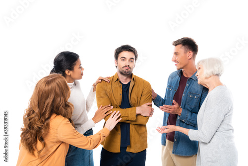 Sad man with crossed arms standing near multiethnic friends isolated on white