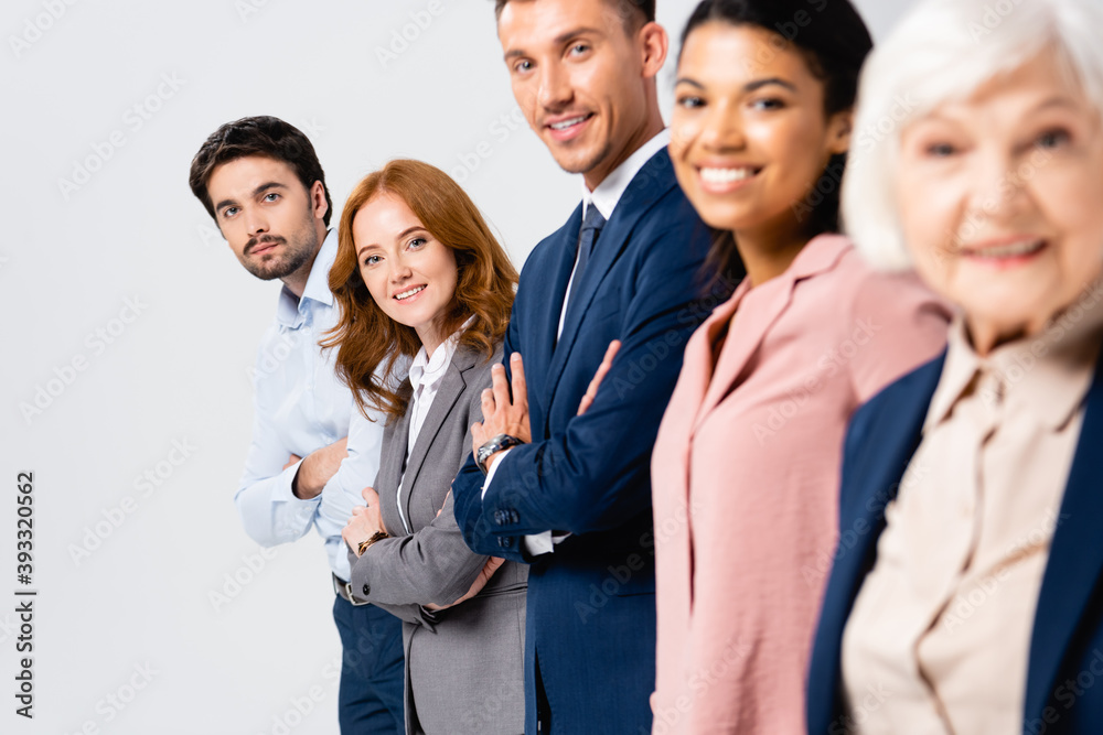 Smiling businesspeople with crossed arms looking at camera near multiethnic colleagues on blurred foreground isolated on grey Smiling businesspeople with crossed arms looking at camera near multiethnic colleagues on blurred foreground isolated on grey