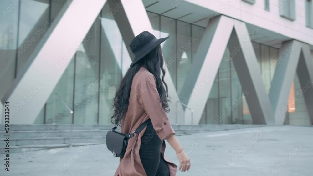 Fashionable brunette young woman wearing a pink coat and a black hat walking to the right on the sidewalk. Slow motion, 4k, tracking shot, side view, cinematic
