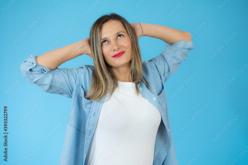 Young woman standing over blue isolated background relaxing and stretching, arms and hands behind head and neck smiling happy Young woman standing over blue isolated background relaxing and stretching, arms and hands behind head and neck smiling happy