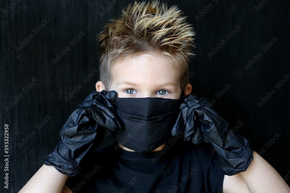 A five-year-old boy wearing a mask and gloves is protected from coronavirus. Blond boy in black medical mask against coronavirus.Boy in black mask on a black isolated background. A five-year-old boy wearing a mask and gloves is protected from coronavirus. Blond boy in black medical mask against coronavirus.Boy in black mask on a black isolated background.