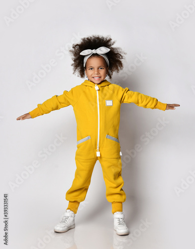 Happy curly dark-skinned kid girl in warm yellow sports jumpsuit and with hair bow stands holding arms up spread