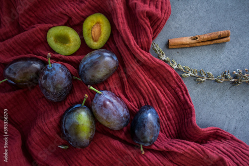Fresh plums, cinnamon, knife and rosemary on a table. Red textured cotton drapery on background.  Fruits still life photo. 