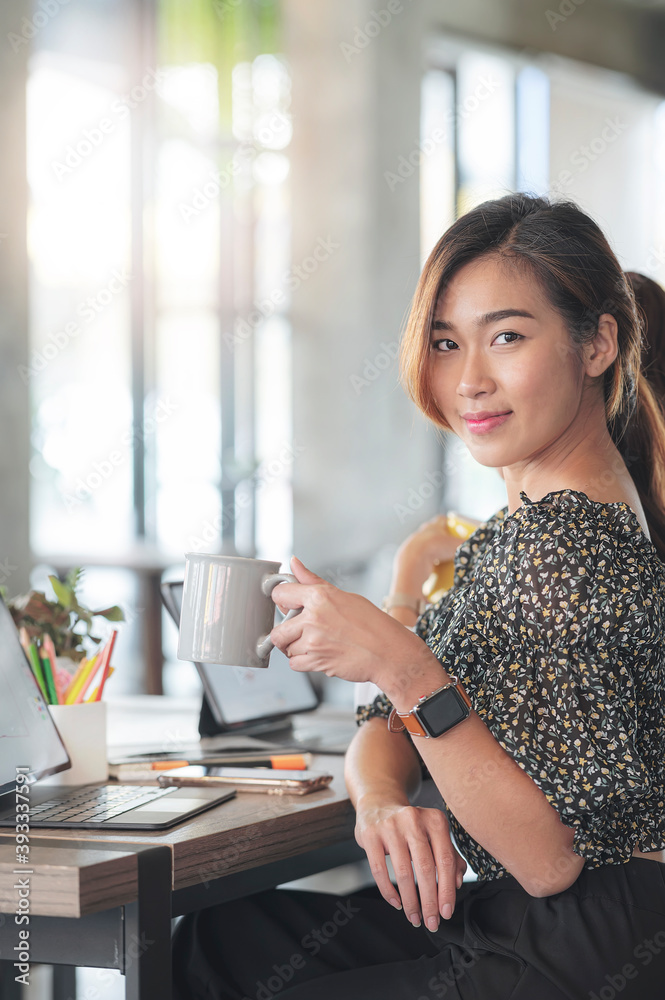 young asian woman holding yellow mug, smiling and looking into camera while sitting at office desk in modern office room. young asian woman holding yellow mug, smiling and looking into camera while sitting at office desk in modern office room.