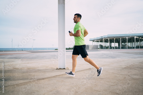 Side view of determined male athlete in electronic headphones listening music songs during cardio running around city, Caucaisan sportsman in modern earphones running during physical recreation