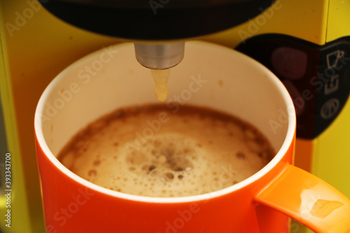 Coffee in a bright orange сup on the table. Close-up is a light green coffee maker with an orange cup.