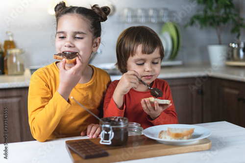 Sister and brother alone in the kitchen have breakfast on their own chocolate paste, smeared on the toast of bread at the table in the kitchen. Children have fun eating chocolate mousse