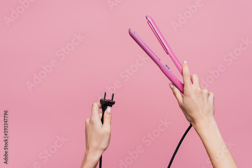 cropped view of woman holding hair straightener in hands isolated on pink