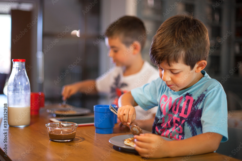 Two caucasian children having breakfast on their own, at daytime in a modern kitchen. Brothers making their own breakfast, pancakes and paste chocolate. Two caucasian children having breakfast on their own, at daytime in a modern kitchen. Brothers making their own breakfast, pancakes and paste chocolate.