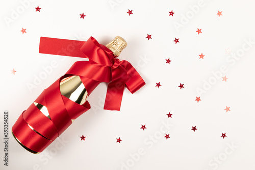 a gift bottle of champagne in golden foil wrapped in a red silk ribbon and scattered confetti on a white background