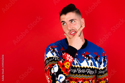 Young handsome Caucasian man wearing Christmas sweater against red wall, Thinking worried about a question, concerned and nervous with hand on chin.