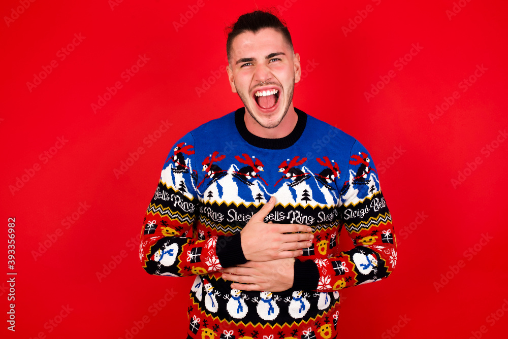 Young handsome Caucasian man wearing Christmas sweater against red wall, smiling and laughing hard out loud because funny crazy joke with hands on body. Young handsome Caucasian man wearing Christmas sweater against red wall, smiling and laughing hard out loud because funny crazy joke with hands on body.