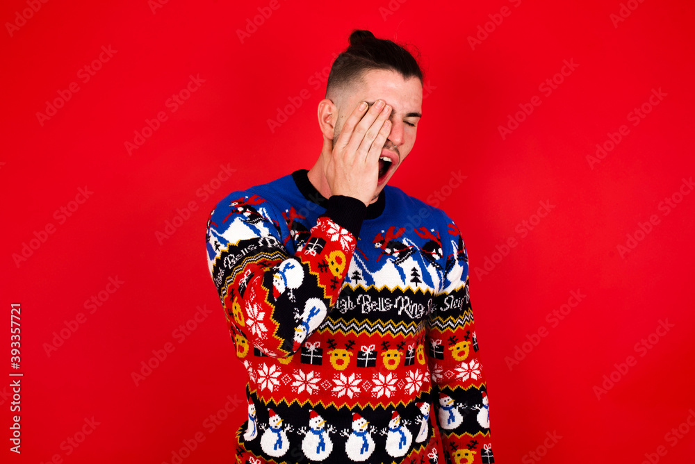 Young handsome Caucasian man wearing Christmas sweater against red wall, Yawning tired covering half face, eye and mouth with hand. Face hurts in pain.