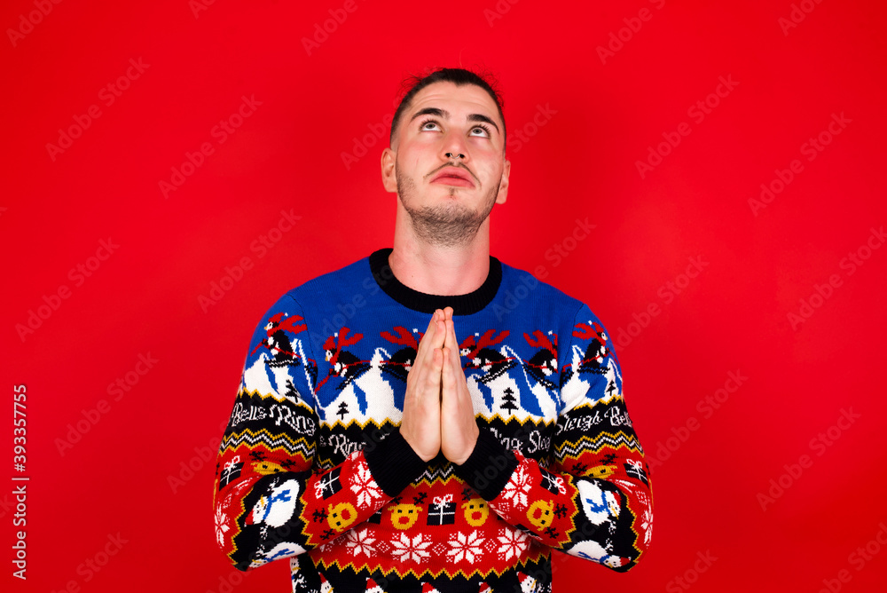 Young handsome Caucasian man wearing Christmas sweater against red wall, begging and praying with hands together with hope expression on face very emotional and worried. Please God Young handsome Caucasian man wearing Christmas sweater against red wall, begging and praying with hands together with hope expression on face very emotional and worried. Please God