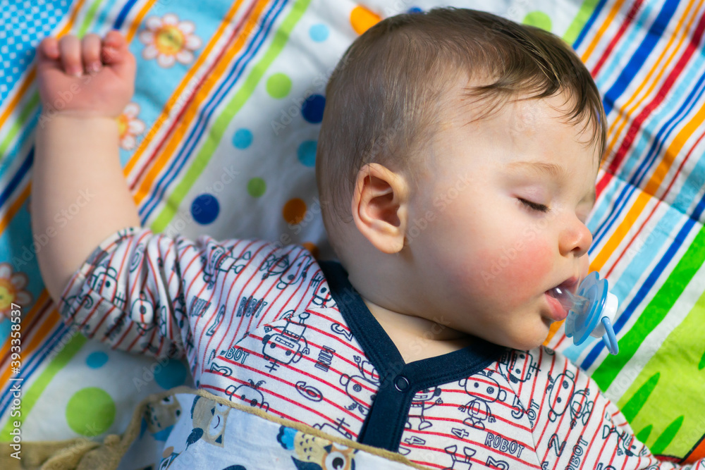 Little caucasian baby boy with red cheeks sleeping, holding his pacifier in mouth on colorful blanket background.