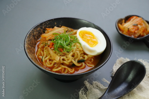 Instan ramyeon or Korean noodles soup on black bowl, topped with half boiled egg, leek and Kimchi on small bowl photo