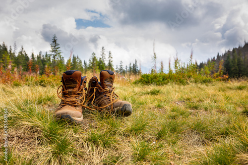 Fototapeta Naklejka Na Ścianę i Meble -  Trekking shoes standing on dry autumn grass on a mountain glade Rowien Waksmundzka with pine trees and spruces in the background, in Tatra Mountains, Poland