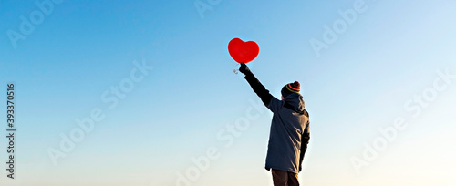 Billede på lærred young man from the back in warm clothes holding a red heart-shaped balloon against the blue sky