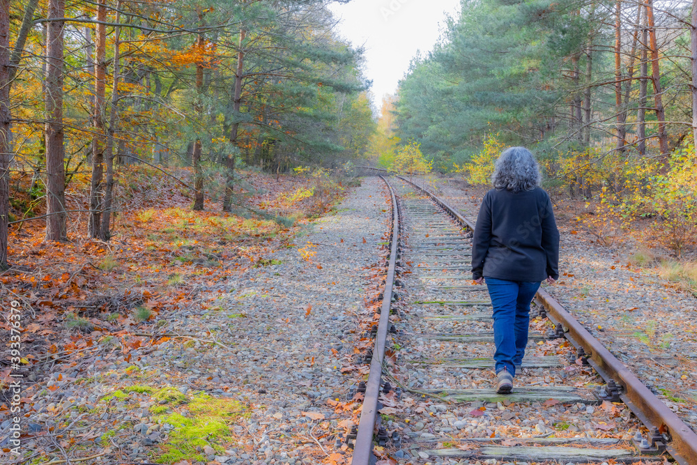Old Rhine Iron Railway (IJzeren Rijn) with a mature woman with grayish black hair walking on train tracks, back to camera, autumnal trees in the Meinweg nature reserve in Middle Limburg, Netherlands
