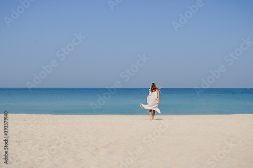 A blonde smiling girl in a maxi linen dress with fluttering hair jumping and dancing on a sand beach against blue sky and sea background.