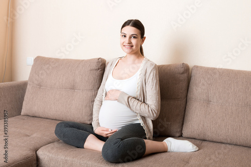 photo of cute caucasian future mom lying in light living room on sofa and holding hands on tummy