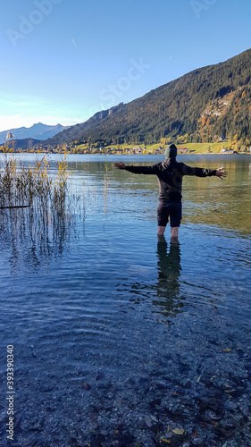 A man standing in the cold water of Weissensee, with the mountains view around. The man spreads his arms wide open in the gesture of freedom. The Austrian Alps are changing colors for winter.