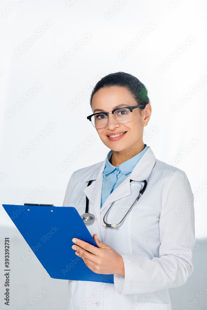 Front view of happy african american female doctor with clipboard looking at camera, while standing on blurred background