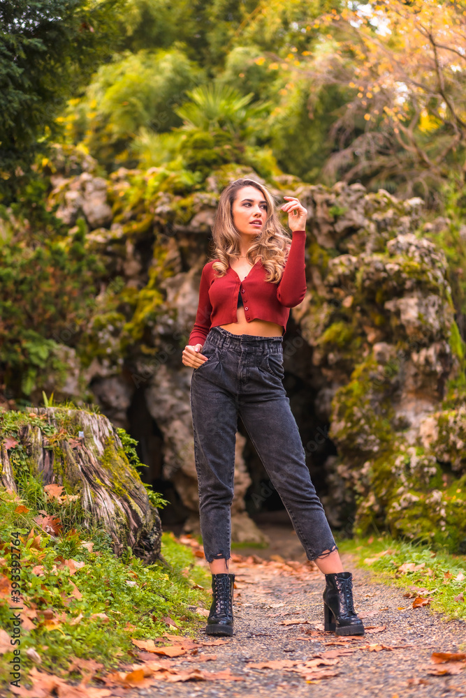 Autumn lifestyle, blonde Caucasian girl in a red sweater and jeans and with high heels, enjoying nature in a park with a natural cave in the background
