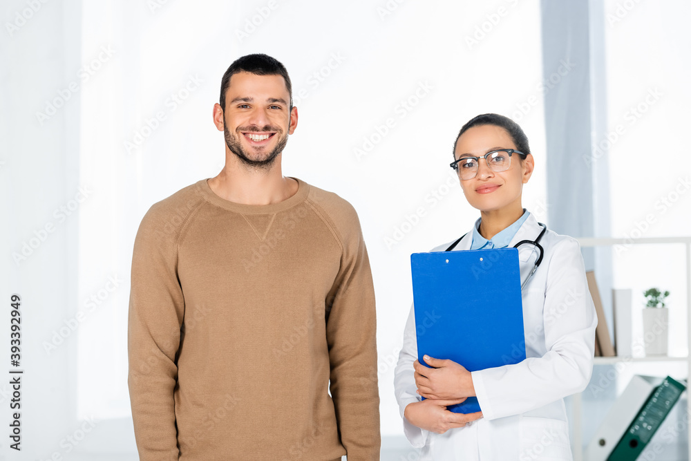 Front view of multicultural patient and doctor standing near each other and looking at camera on blurred background Front view of multicultural patient and doctor standing near each other and looking at camera on blurred background