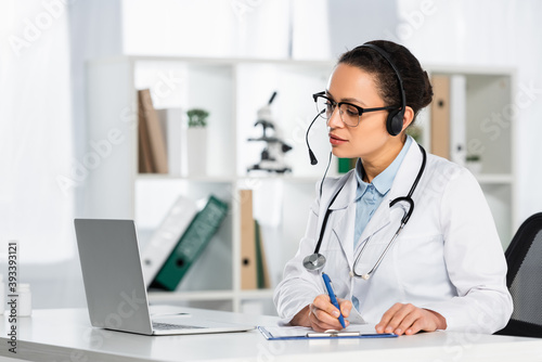 African american doctor in headset writing on clipboard, while looking at laptop during online consultation on blurred background