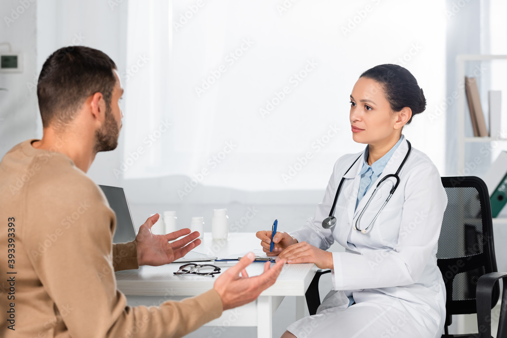 African american doctor looking at patient gesturing, while writing on clipboard at workplace on blurred background African american doctor looking at patient gesturing, while writing on clipboard at workplace on blurred background