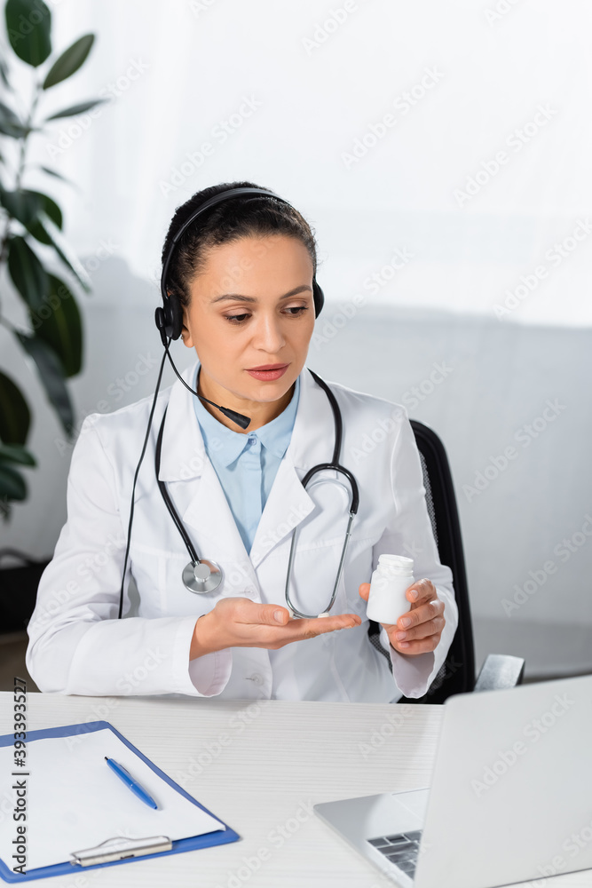 African american doctor in headset pointing with hand at bottle with pills, while looking at laptop on blurred background African american doctor in headset pointing with hand at bottle with pills, while looking at laptop on blurred background