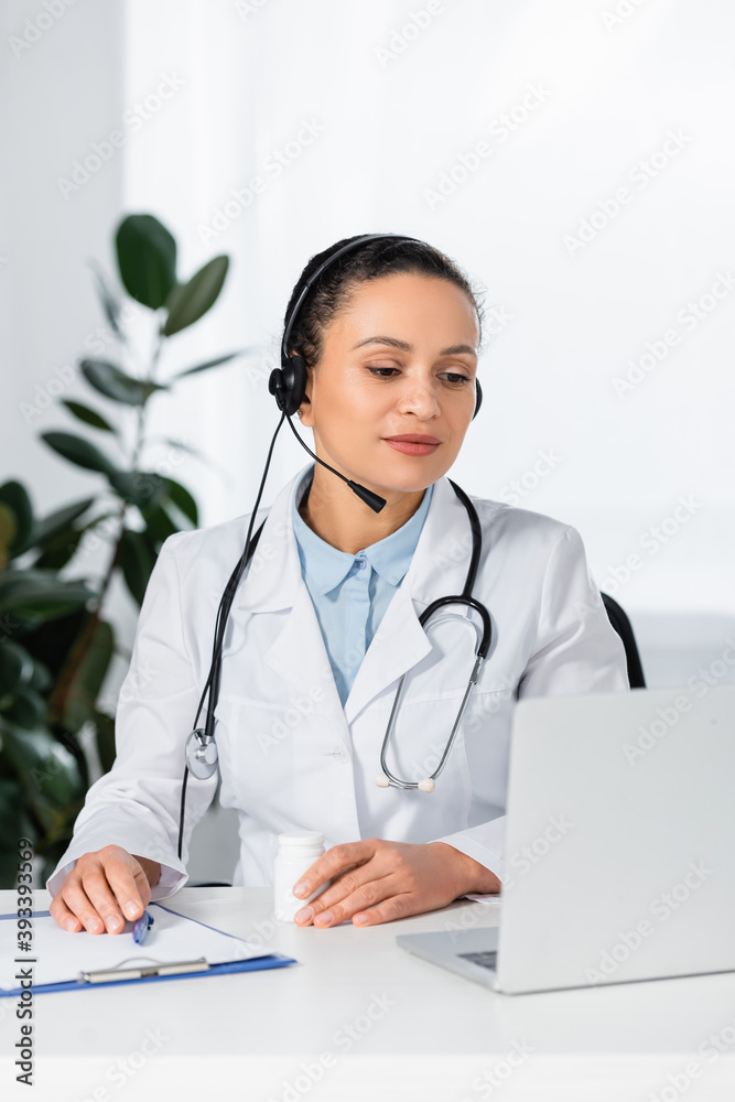 African american doctor in headset looking at laptop, while sitting at workplace on blurred background African american doctor in headset looking at laptop, while sitting at workplace on blurred background