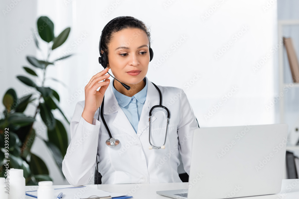 African american doctor in headset looking at laptop sitting near bottles with pills at workplace on blurred background African american doctor in headset looking at laptop sitting near bottles with pills at workplace on blurred background