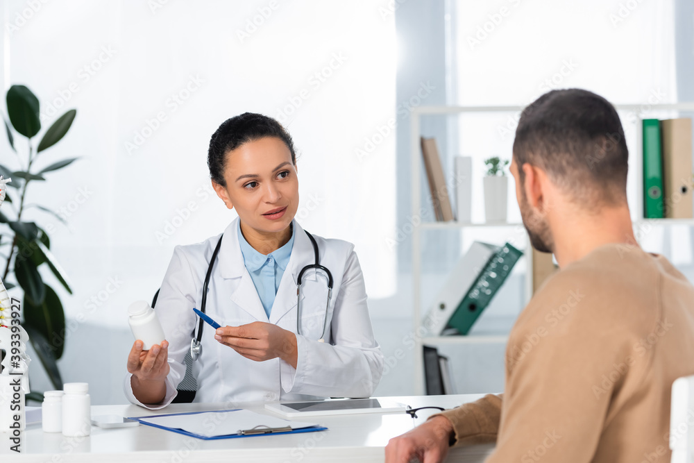 African american doctor pointing with pen at bottle with pills, while looking at patient at workplace on blurred background