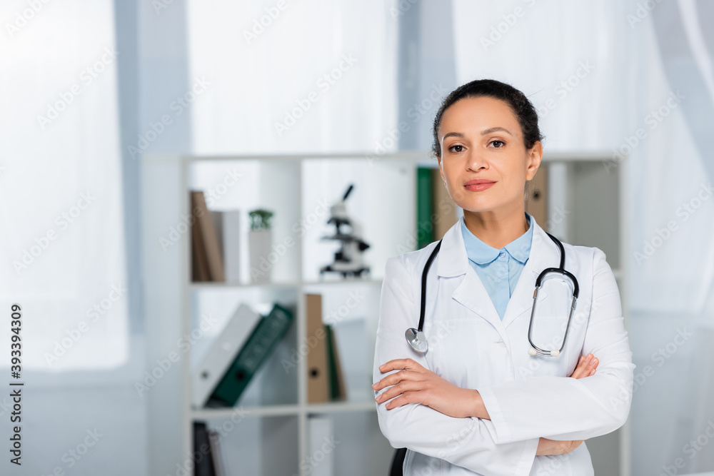 Front view of confident african american doctor with crossed arms looking at camera in clinic on blurred background Front view of confident african american doctor with crossed arms looking at camera in clinic on blurred background