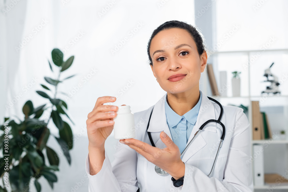 Positive african american doctor showing bottle with pills with blurred clinic on background Positive african american doctor showing bottle with pills with blurred clinic on background
