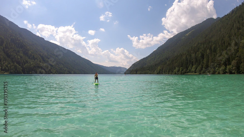 A man stand-up paddling on the crystal clear water of Weissensee lake in Austrian Alps. The lake is surrounded by high mountains. Exercising in the nature. Serenity and calmness. © Chris