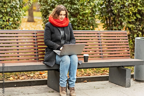 Young woman working on laptop sitting on a park bench with coffee. Computer and online shopping use. Distance learning, online education.