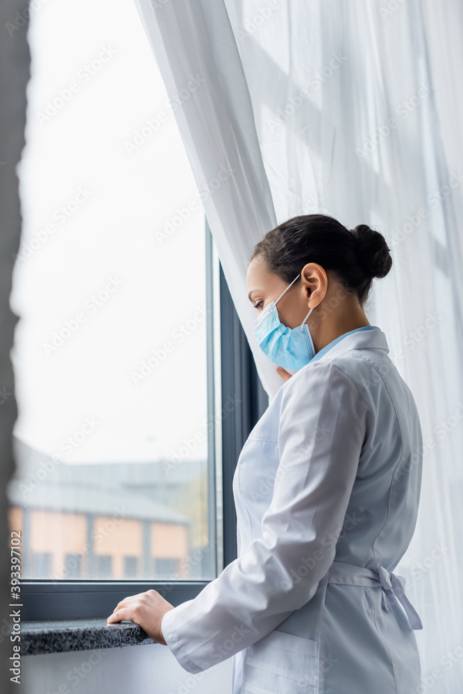 Upset african american doctor in medical mask standing near window in clinic Upset african american doctor in medical mask standing near window in clinic