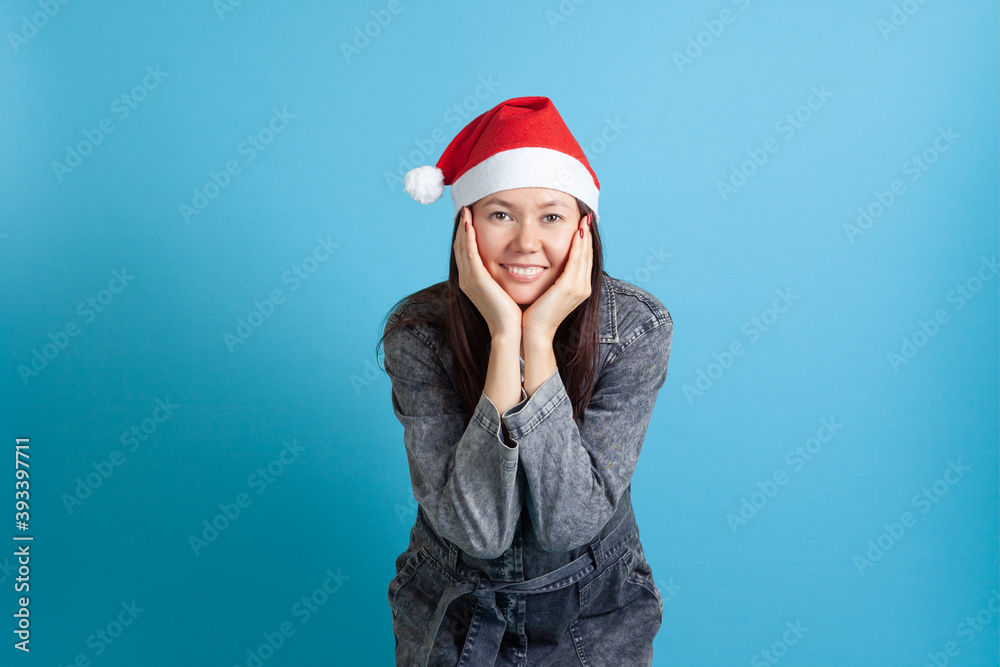 mock up portrait of a smiling Asian young woman in a Santa hat with her head in her hands, on a blue background. mock up portrait of a smiling Asian young woman in a Santa hat with her head in her hands, on a blue background.