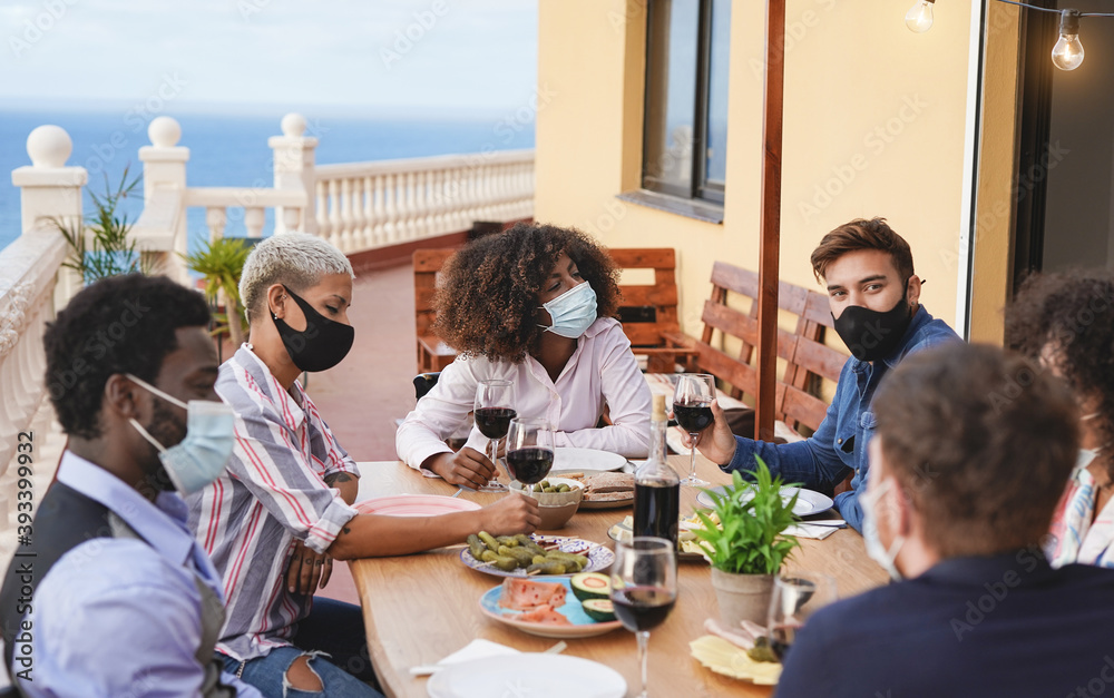 Obraz premium Cheerful young multiracial friends enjoy dinner at home on terrace while wearing protective face mask for coronavirus