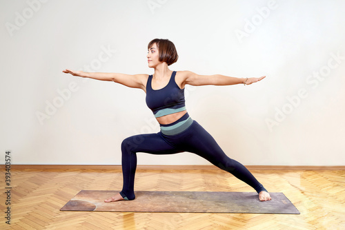 Brunette woman in navy blue yoga tights giving yoga poses in front of white wall. The woman in quarantine due to Covid-19 exercises alone. Brunette woman doing sports routinely in quarantine. photo